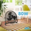 A black floor fan is placed on a wooden floor in a room with a window and a desk with a chair. Voir les images du produit