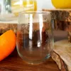 A close-up of a stemless wine glass filled with a dark liquid and ice, placed on a wooden surface next to a slice of orange. The glass has condensation on the outside. In the background, there are blurred out drinks and wooden stands. Produktbild-Vorschau