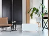 A modern living room with a white electric glass heater in the foreground, a leather chair and a small table to the left, and a large potted plant to the right. Produktbild-Vorschau