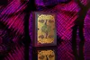 A deck of Bicycle playing cards with a purple and gold peacock design is displayed on a reflective black surface, with a blurred purple and pink background. Produktbild-Vorschau