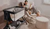 A woman reads a book while sitting in a rocking chair next to a baby crib. Produktbild-Vorschau
