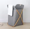 A grey fabric laundry basket with wooden supports and a white towel draped over the edge, placed on a wooden floor against a white wall. Náhľad obrázku produktu