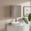 A modern bathroom vanity with a sink, faucet, and a mirrored cabinet above. A framed picture and a large plant are visible on the right. Náhľad obrázku produktu