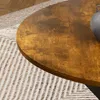 A close-up view of a round wooden table with a textured surface, placed on a rug with a geometric pattern. A dark gray vase is partially visible on the right side of the table. Produktbild-Vorschau
