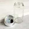A clear glass spice jar with its silver lid, which has holes for dispensing spices, is shown on a light-colored wooden surface. Produktbild-Vorschau
