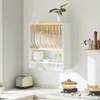 A white kitchen wall-mounted dish rack with wooden dividers, holding white plates. Below are white bowls, glasses, and mugs hanging from hooks. A toaster is on the counter to the left, and a stovetop with pots is to the right. Produktbild-Vorschau