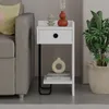 A modern white nightstand with a drawer and shelf, placed next to a gray sofa and a potted plant. Produktbild-Vorschau