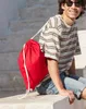 A young man wearing a striped t-shirt and denim shorts is sitting on steps with a red drawstring bag over his shoulder. Produktbild-Vorschau