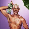 A shirtless man with blonde hair holds a bottle of Jean Paul Gaultier Le Beau Paradise Garden perfume on his shoulder, smiling at the camera. A large green leaf is visible in the upper right corner. Anteprima dell’immagine prodotto