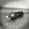 A corded rotary tool with a grinding attachment rests on a textured metallic surface, with a brick wall in the background. Produktbild-Vorschau