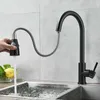 A black kitchen faucet with a pull-down sprayer is shown in use, with water flowing into a stainless steel sink. A hand is holding the sprayer. Produktbild-Vorschau