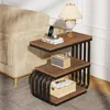 A 3-tier side table with a vintage industrial design, placed next to a sofa in a living room setting. The table holds a lamp, a tissue box, books, and a phone charging. Produktbild-Vorschau