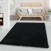 A black shaggy rug is placed on a light-colored floor in a living room setting. A modern wooden sofa with light-colored cushions is visible in the background. Produktbild-Vorschau