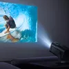 A mini projector is projecting an image of a surfer on a wave onto a wall. The projector is placed on a dark table next to a couch. Voir les images du produit
