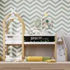 A children's bunk bed with a house-shaped frame, a chalkboard front, and stairs with drawers, set against a chevron-patterned wall. Produktbild-Vorschau
