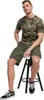 A man is sitting on a stool wearing a camouflage t-shirt and olive green shorts. Náhled obrázku produktu