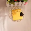 A heart-shaped chocolate sits on a gold wrapper, which is placed on a doily. In the background, there is a white fence with flowers. Produktbild-Vorschau