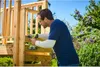 A man is using a cordless drill to assemble a wooden structure, possibly a treehouse or playhouse, outdoors on a sunny day. Produktbild-Vorschau