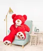 A large red teddy bear is sitting in a light blue armchair in a room with a floor lamp and small tables. Produktbild-Vorschau