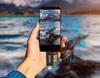 A hand holding a smartphone with a USB-C card reader attached, showing a scenic view on the phone screen. Icons indicating data transfer are visible. Produktbild-Vorschau