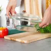 A person is holding a glass baking dish over a wooden cutting board, with an apple and parsley in the foreground. Podgląd zdjęcia produktu