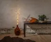 A decorative twig with LED lights in a brown vase, placed on a wooden floor next to a bench with a plant and a decorative carrot. Podgląd zdjęcia produktu