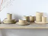 A collection of ceramic dishes and cups arranged on a wooden shelf against a white wall. Produktbild-Vorschau