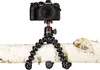 A black Olympus camera is mounted on a red and black Joby GorillaPod tripod, which is wrapped around a piece of white birch wood. Produktbild-Vorschau