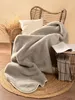A cozy scene with a gray wool blanket draped over a wicker chair, next to a small side table with a book. Produktbild-Vorschau