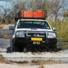 A white off-road vehicle driving through shallow water, with orange recovery tracks on its roof rack. Náhled obrázku produktu