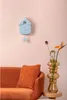 A light blue cuckoo clock is mounted on a pink wall above an orange sofa with an orange textured pillow. A vase with flowers is on a side table to the right. Náhled obrázku produktu
