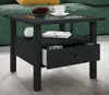 A black side table with a glass top, a shelf, and an open drawer, placed in a living room setting with a green sofa and a yellow cushion. Voir les images du produit