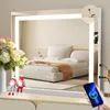 A bedroom vanity with a large illuminated mirror reflecting a bed. A phone is being charged via a USB port on the mirror.