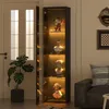 A tall, narrow glass display cabinet with LED lighting, showcasing various decorative figurines, is placed next to a door and a large potted plant. A sofa is visible in the background. Náhľad obrázku produktu