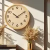 A wooden wall clock with black numbers and hands is displayed on a white wall, with shadows cast by sunlight. Dried grass in a golden vase is placed in the foreground. Produktbild-Vorschau