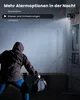 A person in a hooded jacket is seen from behind, holding a bag and approaching a door. A security camera is mounted on the wall above the door, emitting a beam of light and red warning signals. Produktbild-Vorschau