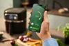 A hand holding a smartphone displaying an air fryer app, with an air fryer and kitchen ingredients in the background. Podgląd zdjęcia produktu