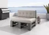 A modern outdoor lounge set made from grey wooden pallets with beige cushions, placed on a concrete patio overlooking the sea. A grill is visible to the left, and a decorative lantern and plant are to the right. Produktbild-Vorschau