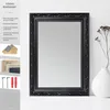 A black ornate mirror is displayed on a white surface next to a white vase with dried flowers. Produktbild-Vorschau