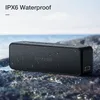 A black portable Bluetooth speaker with water droplets on it, placed on rocks near the water. Produktbild-Vorschau