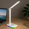 A white LED desk lamp with a colorful RGB base is placed on a wooden desk next to a laptop and a notebook. Produktbild-Vorschau