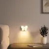 A butterfly-shaped LED night light is mounted on a wall above a wooden table, casting a warm glow. A potted plant and glasses are on the table. Produktbild-Vorschau
