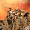 A detailed wooden model of a steam locomotive is positioned on a track, with dry grass and rocky terrain in the foreground and a dramatic orange sky in the background. Náhľad obrázku produktu