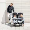 A man stands next to a double stroller with two children in it, outdoors. Produktbild-Vorschau