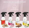 Four spray bottles with different labels and scents, along with decorative elements like cookies, flowers, and wood sticks, are displayed against a soft pink background. The image also features text and a logo related to fragrances. Produktbild-Vorschau