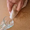 A hand is wiping a spill off a cork surface next to a glass. Náhľad obrázku produktu