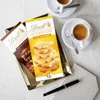 Two Lindt Les Grandes chocolate bars are displayed on a gold tray next to two cups of coffee and a pen on a patterned napkin. Produktbild-Vorschau