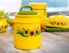 A close-up shot of a yellow ceramic jar with a green lid, decorated with sunflowers. Produktbild-Vorschau