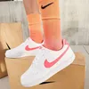 A person wearing white Nike sneakers with pink accents and orange socks with a black Nike logo, standing on cardboard boxes. Produktbild-Vorschau