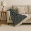 A dark gray herringbone patterned throw blanket with beige fringe is draped over the arm of a light beige sofa. A matching beige pillow is on the sofa, and a light beige lamp sits on a small wooden side table next to the sofa. A light beige rug is on the f Anteprima dell’immagine prodotto